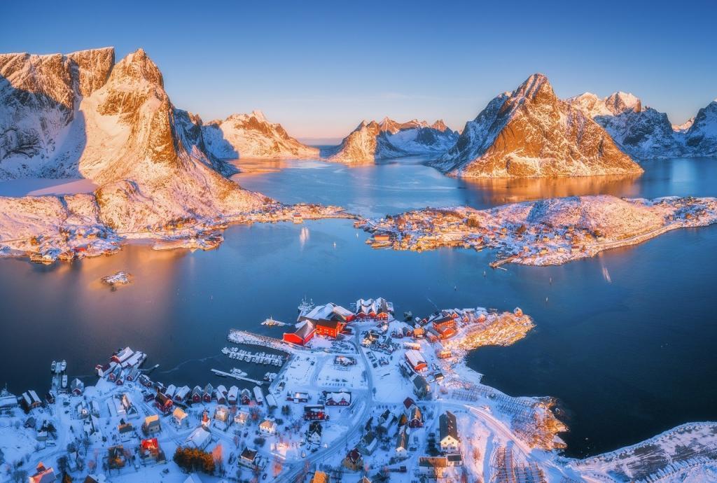Aerial view of Reine at sunrise in winter. Top view of Lofoten islands, Norway. Panoramic landscape with blue sea, snowy mountains, high rocks, village, buildings, rorbu, sky reflected in water