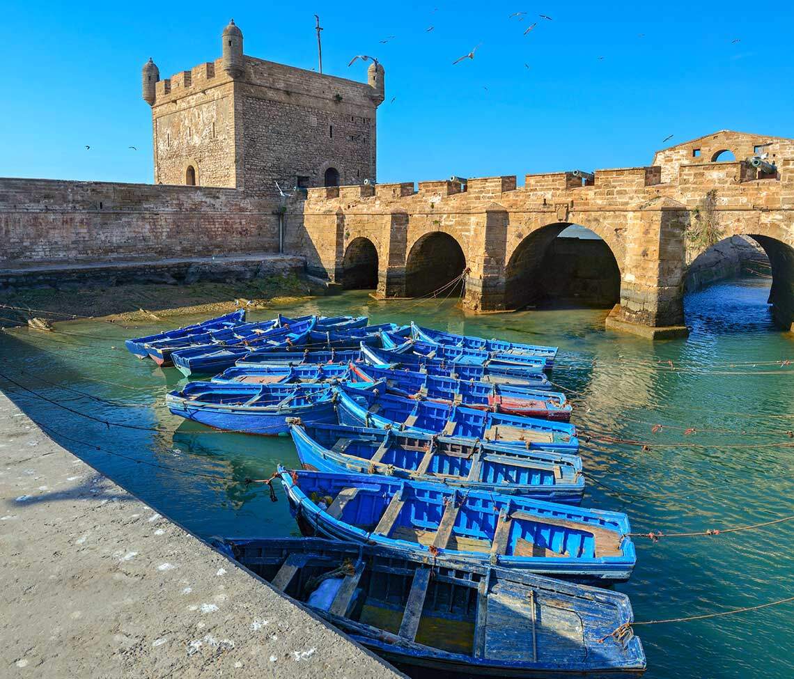 Pemandangan pelabuhan Essaouira dengan kapal nelayan biru dan kubu Castelo Real of Mogador