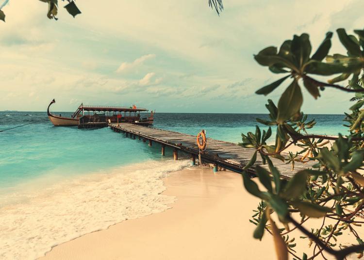 View of a wooden pier leading to a beautiful gondola moored at the end of it, surrounded by teal ocean water; coral sand of a Maldivian island with greenery, and a wave foam in the foreground